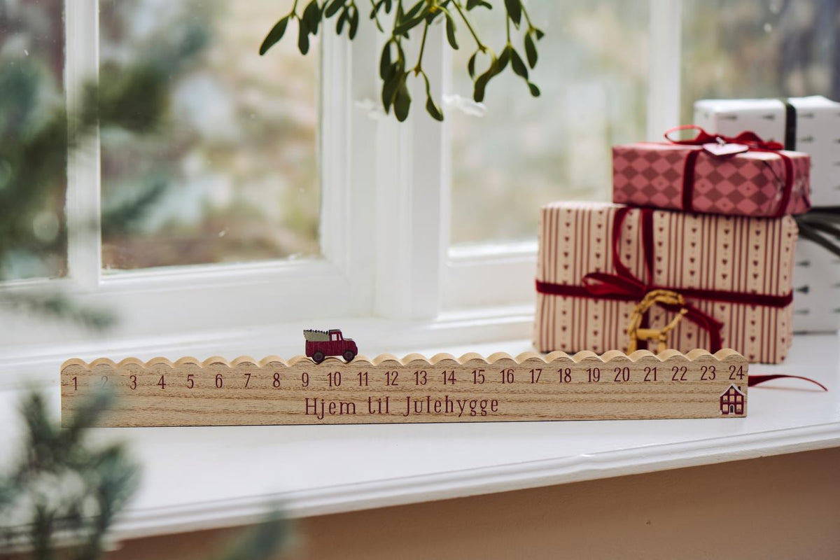 Julekalender i MDF med rød lastbil og juletræ, hvor bilen flyttes fra 1-24 mod huset – køb hos butikalrum.dk.